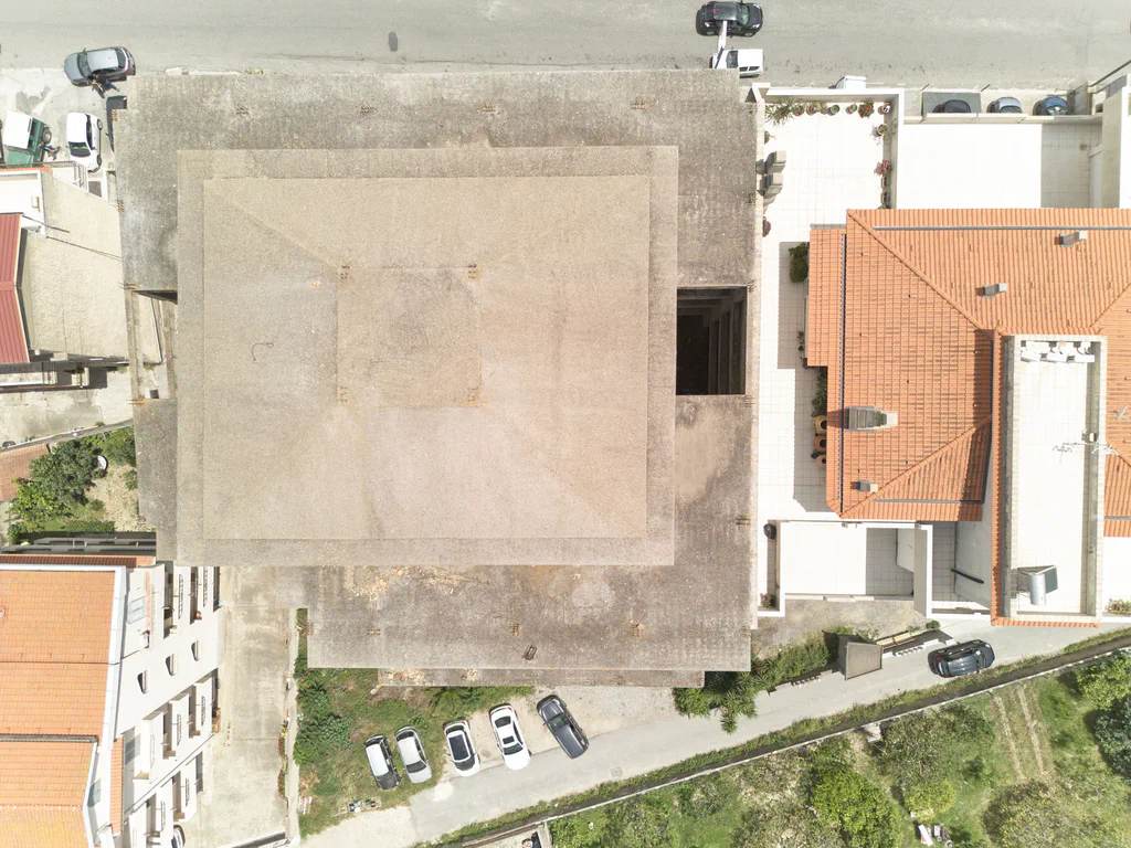 Aerial view of a flat-roofed concrete building among tiled roofs, a road and parked cars