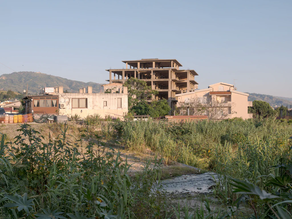 Unfinished structure on a rise seen across an overgrown field, mountains behind