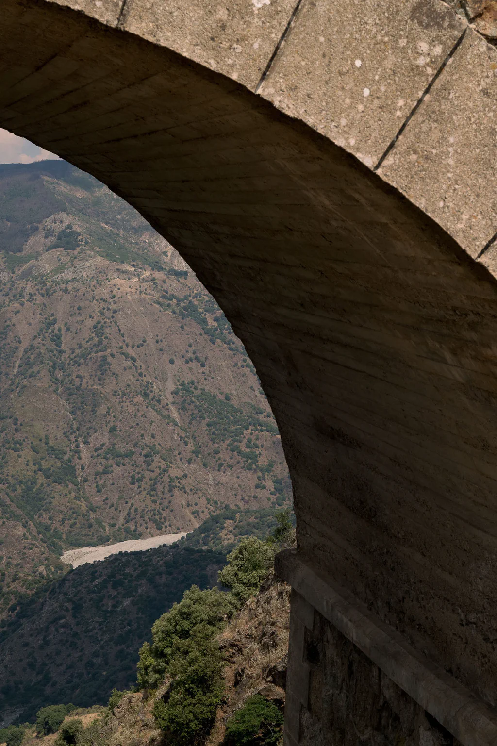 Stone arch of an old bridge framing a wooded valley and a road far below
