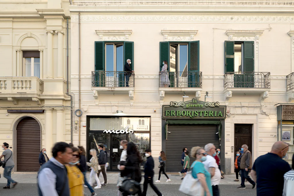 Passers-by in front of a herbalist’s shop, below a pale façade with green shutters