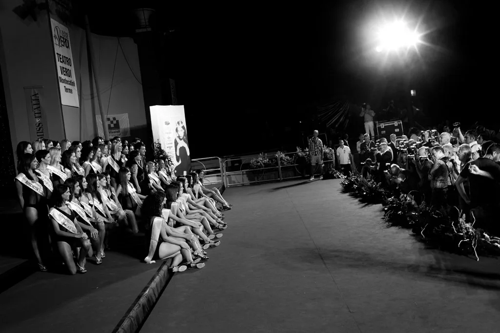 Contestants seated along the edge of a stage under a bright spotlight