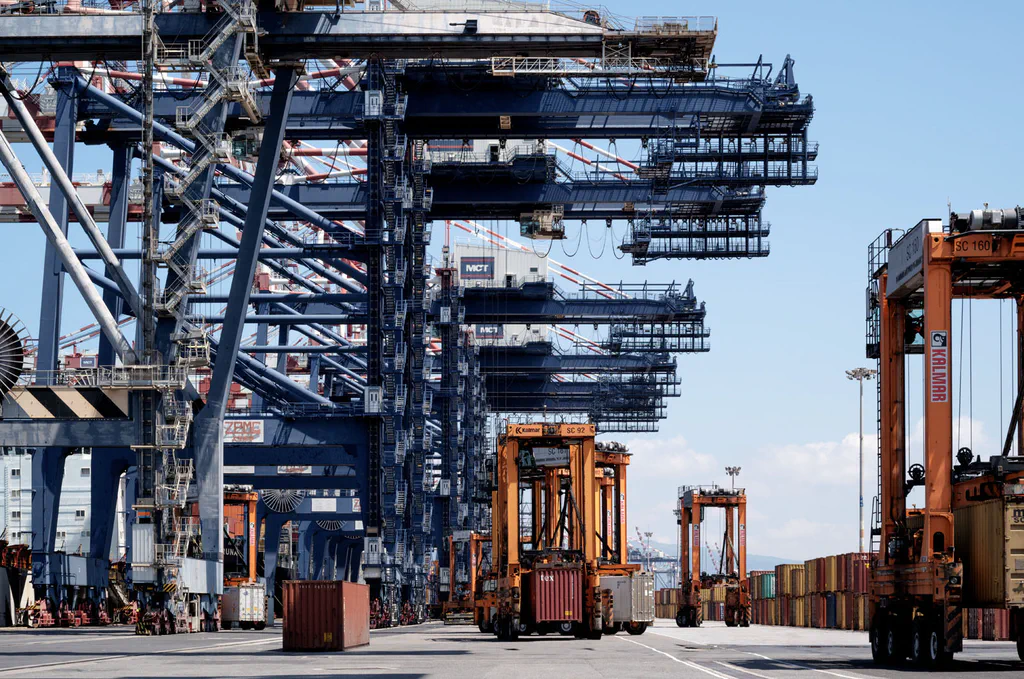 Row of blue gantry cranes seen end on, a straddle carrier below