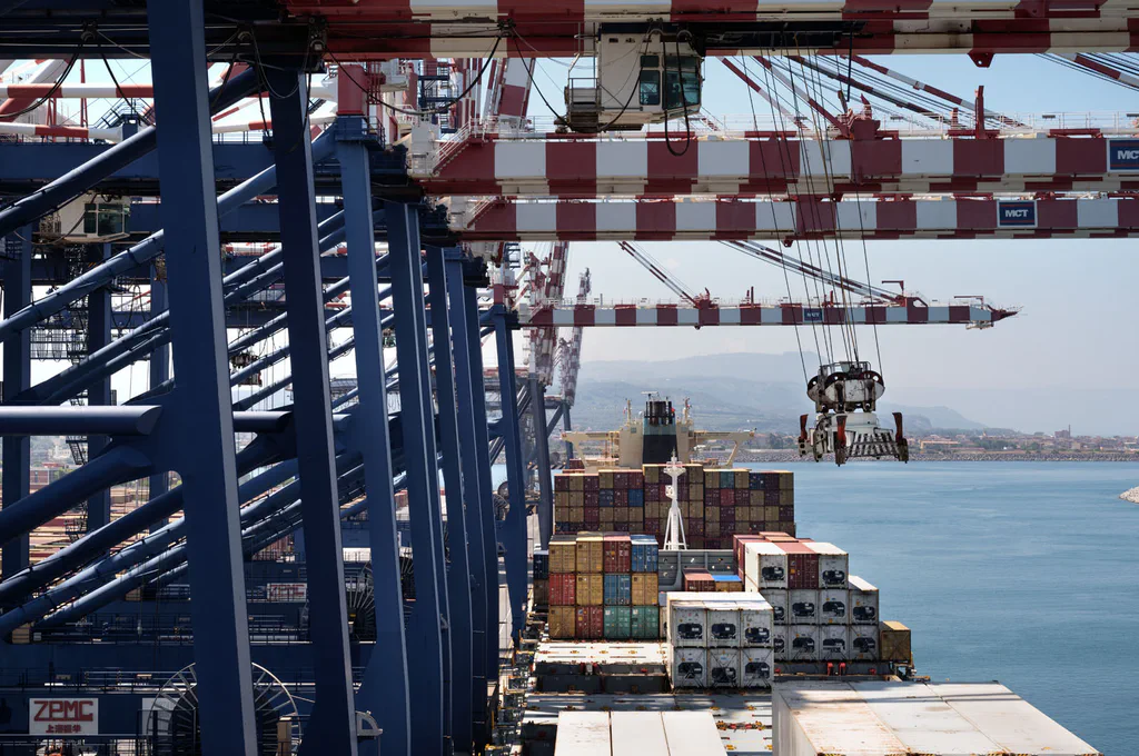 Cranes over the water, the coastline visible beyond the containers