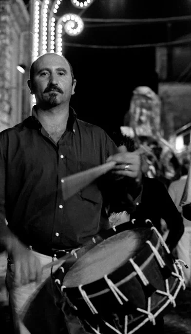 Man playing a large drum at night, festival lights on the building behind