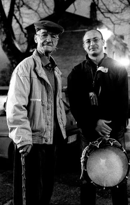 Two men standing side by side at night, one holding a drum