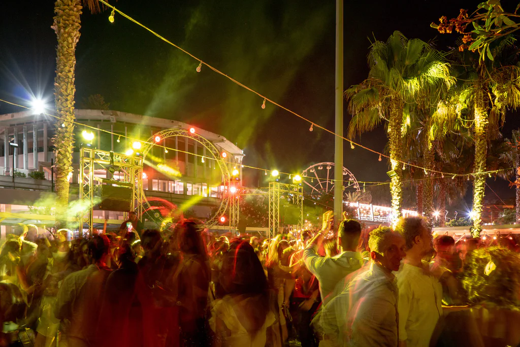 A night crowd among palms and coloured lights on the seafront