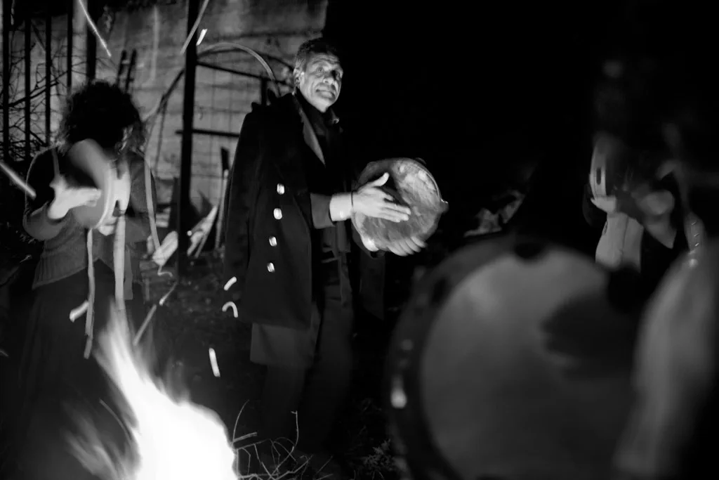 Man playing a frame drum by firelight at night, dancers around him