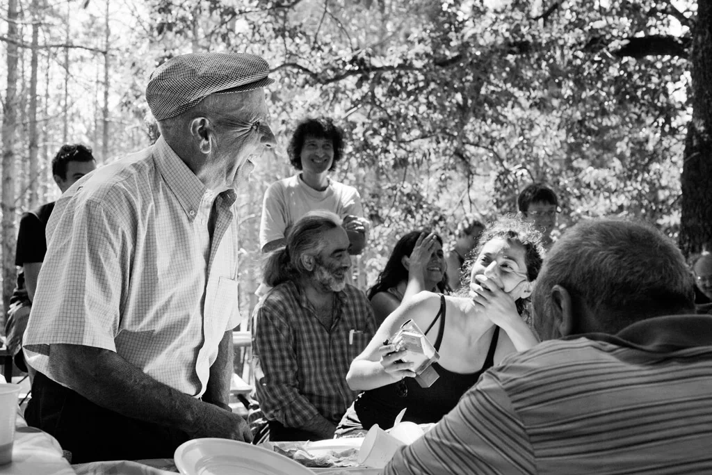 Elderly man in a flat cap playing a wind instrument outdoors, people listening on the grass
