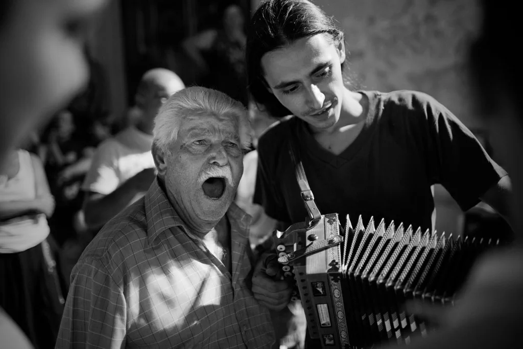 Elderly man singing beside a young accordion player at a night gathering