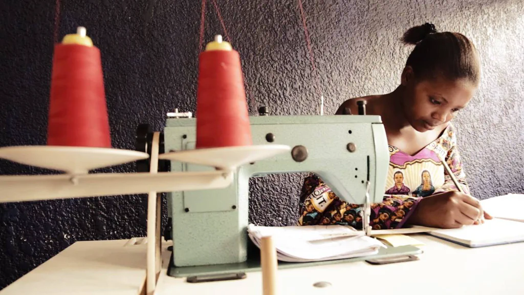 A young woman writing at a table beside an industrial sewing machine with two cones of red thread