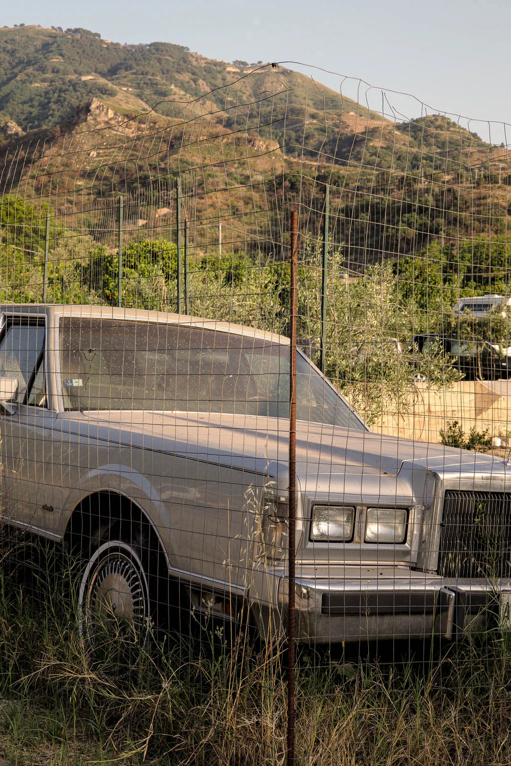 Abandoned car behind a wire fence, wooded hills rising behind